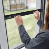 En person i grå ternet skjorte installerer et Myggenet til vindue - Rullegardin på en hvid vinduesramme med græs udenfor.