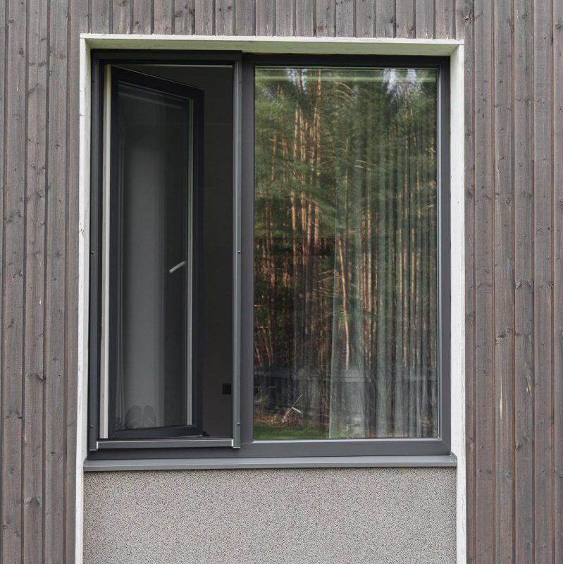 Ein teilweise geöffnetes Fliegengitterfenster - Rollo in einer Holzwand, Bäume spiegeln sich im Glas.