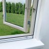 Et åbent vindue med Myggenet til vindue - Rullegardin og en metallås har udsigt til en grøn græsplæne og høje buske.