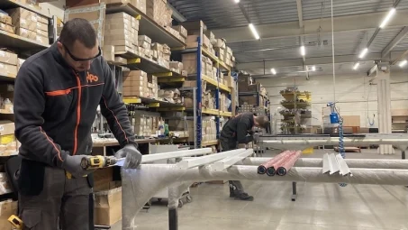 Two workers assemble metal parts at a workstation as Insectnets.com Roll-Up Insect Screen boxes line the warehouse shelves.