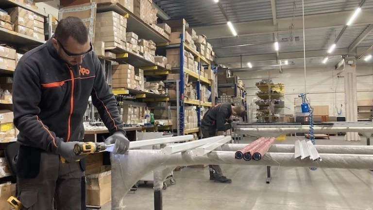 Two workers assemble metal parts at a workstation as Insectnets.com Roll-Up Insect Screen boxes line the warehouse shelves.