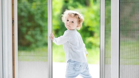 A young child stands at an open sliding screen door with an insect screen, looking back over their shoulder, greenery visible outside.