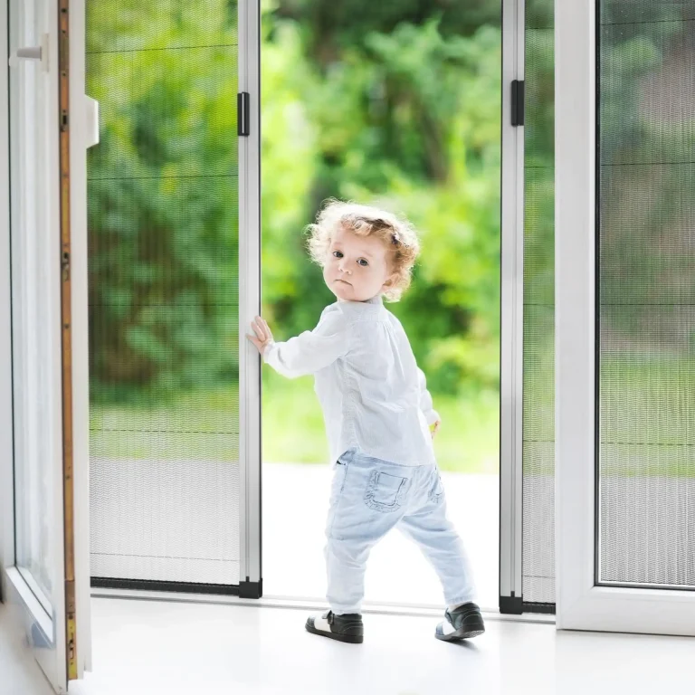 A young child stands at an open sliding screen door with an insect screen, looking back over their shoulder, greenery visible outside.