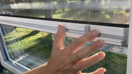 A hand with a ring slides up an Insectnets.com Roll-Up Insect Screen latch, revealing trees and grass outside the home.