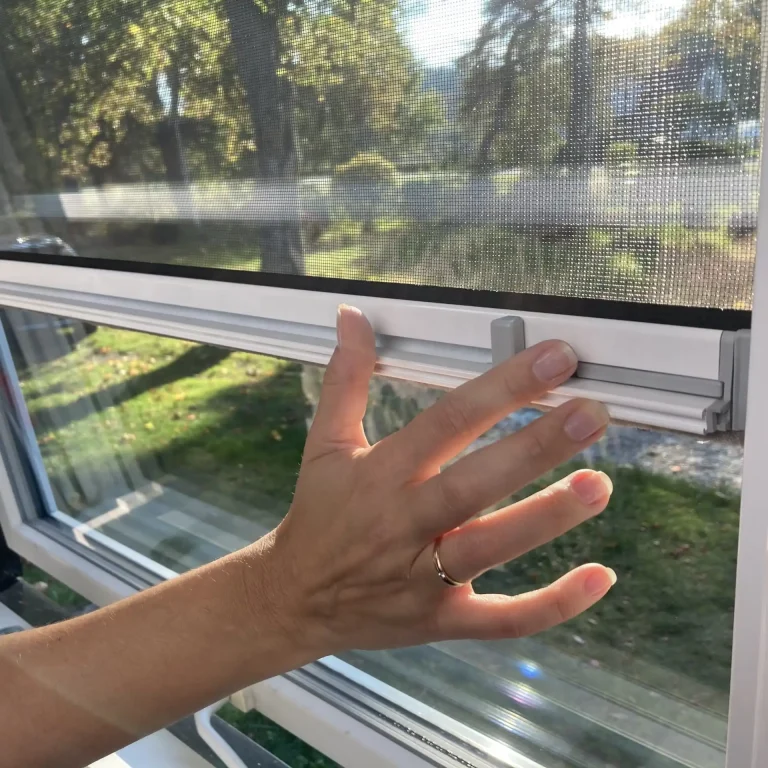 A hand with a ring slides up an Insectnets.com Roll-Up Insect Screen latch, revealing trees and grass outside the home.