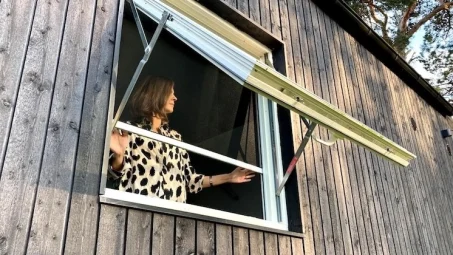 A woman in a patterned shirt opens an awning window with a Roll-Up Insect Screen on the wooden exterior wall of her home.
